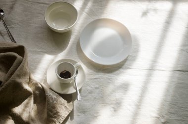 Masa ayarı yakın çekim. Beyaz masa örtüsü üzerine çatal bıçak takımı ve keten peçeteyle boş beyaz tabağın üst görüntüsü. Güneşin altında sakin sakin bir yemek..