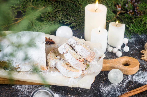 Stollen sliced on kraft paper on dark wooden table with xmas tree branches and decorations,. Christmas German traditional fruit bread stollen with dried berries, nuts and powdered sugar.