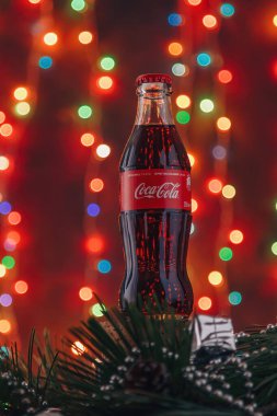 İzmail, Ukrayna. Aralık 2020. Cam Coca Cola şişesi aydınlık bulanık mutlu noeller çam dallarıyla süslenmiş çelenk ışıkları. Dikey çekim
