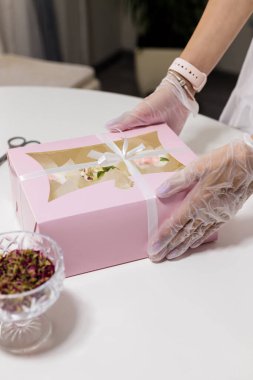 Elleri eldivenli, pembe kek kutusunu tutan şekerci. Masanın üzerinde kurumuş gül yaprakları vazosu. Anneler Günü, Sevgililer Günü, Doğum günü