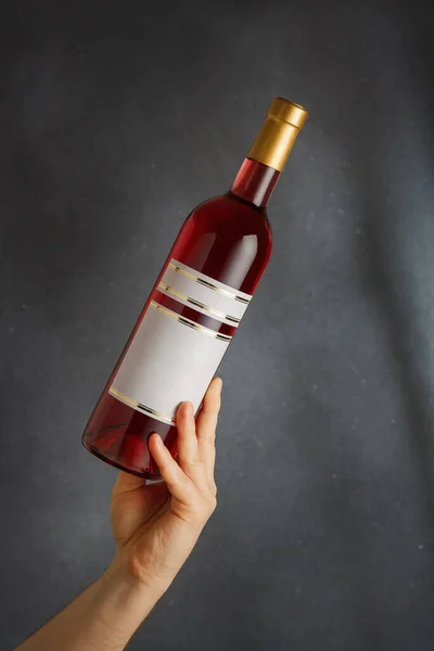 Male hand holding bottle of pink red wine, natural fruit alcohol liquor with empty label on grey background. Mock up, template for brand, vertical shot