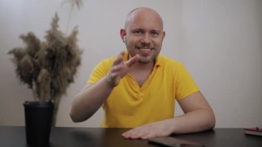 Sarı tişörtlü, kablosuz kulaklıklı, yüzünde piercing olan, kameraya gülümseyen, arkadaşlarıyla veya akrabalarıyla görüntülü konuşan pozitif bir genç.