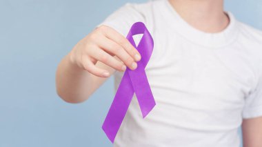 child holding a cancer ribbon on a blue background. purple ribbon in hands.