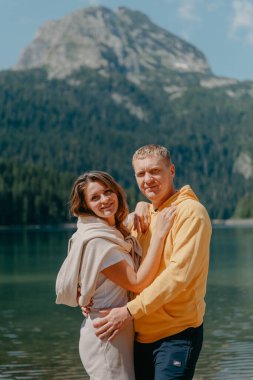 Güzel bir dağ ve gölün kıyısında kucaklaşan çift. Nazikçe sarılıp birbirine aşık çiftin güzel bir doğa geçmişine sahip öpücüğü. Kayalık dağların önünde göl.