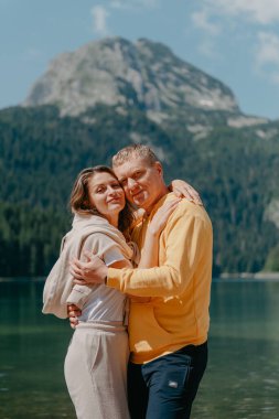 Güzel bir dağ ve gölün kıyısında kucaklaşan çift. Nazikçe sarılıp birbirine aşık çiftin güzel bir doğa geçmişine sahip öpücüğü. Kayalık dağların önünde göl.