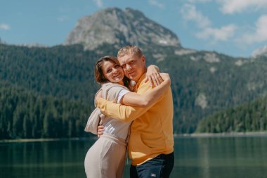 Güzel bir dağ ve gölün kıyısında kucaklaşan çift. Nazikçe sarılıp birbirine aşık çiftin güzel bir doğa geçmişine sahip öpücüğü. Kayalık dağların önünde göl.