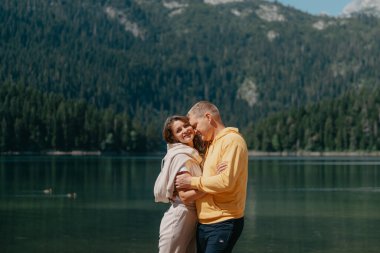 Güzel bir dağ ve gölün kıyısında kucaklaşan çift. Nazikçe sarılıp birbirine aşık çiftin güzel bir doğa geçmişine sahip öpücüğü. Kayalık dağların önünde göl.