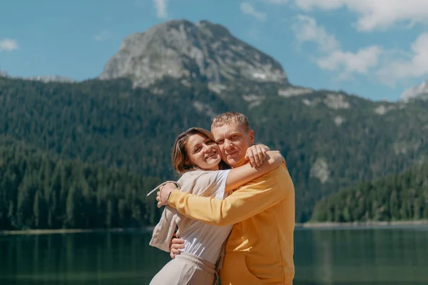 Ζευγάρι αγκαλιάζει στην ακτή μιας όμορφης βουνά και λίμνη. απαλές αγκαλιές και φιλιά του ερωτευμένου ζευγαριού σε ένα όμορφο φόντο της φύσης. λίμνη μπροστά από βραχώδη βουνά. — Φωτογραφία Αρχείου