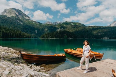 Kız, Karadağ Durmitor Ulusal Parkı 'ndaki balıkçı tekneleri, ormanlar ve dağlarla Kara Göl' e bakıyor. Bir turist sihirli bir göl manzarası, kozalaklı bir orman ve büyülü bir manzaranın tadını çıkarır.