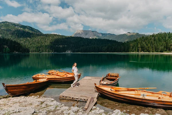 La niña mira el Lago Negro con barcos de pesca, bosques y montañas en el Parque Nacional Durmitor de Montenegro. Un turista disfruta de una vista mágica del lago, un bosque de coníferas y un mágico — Foto de Stock