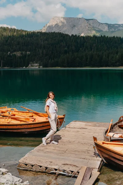 La niña mira el Lago Negro con barcos de pesca, bosques y montañas en el Parque Nacional Durmitor de Montenegro. Un turista disfruta de una vista mágica del lago, un bosque de coníferas y un mágico — Foto de Stock