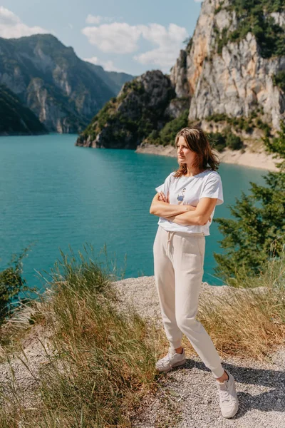Chica turística se encuentra en la orilla de un lago en las montañas. hermoso paisaje, Lago Piva en Montenegro.. Mujer está de pie en la costa del lago Piva al atardecer en verano. Paisaje con chica, famoso — Foto de Stock