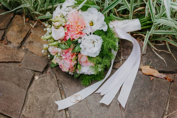 WEDDING GOLD RINGS DAN BRIDAL BOUQUET OF FLOWERS ON STONE BACKGROUND. Buket pernikahan mawar putih dan merah muda dan sepasang cincin pernikahan di atasnya — Stok Foto