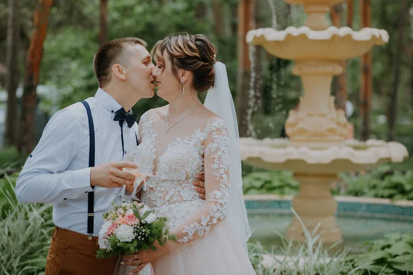 Noiva e noivo com copos de champanhe no parque celebrar o seu dia de casamento. Um casal amoroso de recém-casados bebe champanhe, abraços e beijos no fundo da fonte. Fechar — Fotografia de Stock