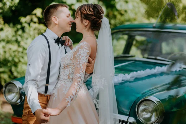 Bara gifta par står nära retro vintage bilen i parken. Sommaren solig dag i skogen. brud i elegant vit klänning med bukett och elegant brudgum i kärlek kramar. Brud och brudgum — Stockfoto