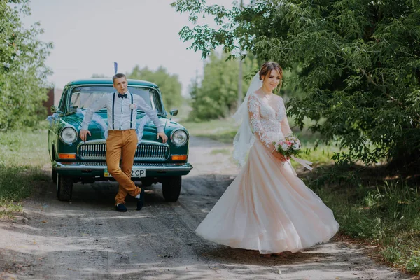 Bara gifta par står nära retro vintage bilen i parken. Sommaren solig dag i skogen. brud i elegant vit klänning med bukett och elegant brudgum. Brudgummen poserar av en — Stockfoto