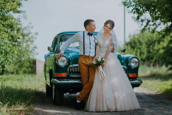 Bara gifta par står nära retro vintage bilen i parken. Sommaren solig dag i skogen. brud i elegant vit klänning med bukett och elegant brudgum i kärlek kramar. Brud och brudgum — Stockfoto