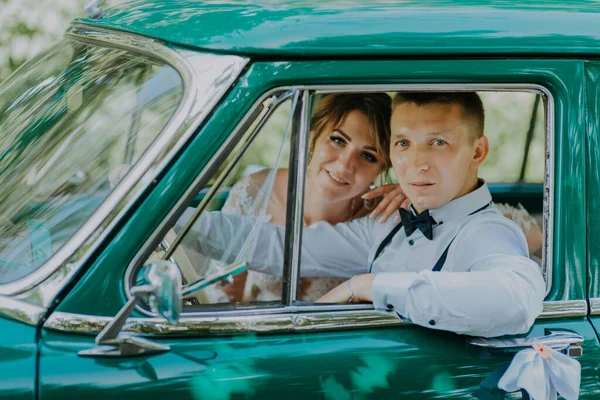 Prachtige stijlvolle blonde bruid poseren in retro groene auto met bruidegom. De bruid en bruidegom zitten in de retro auto. Bruiloft, bruid en bruidegom met een vintage auto — Stockfoto