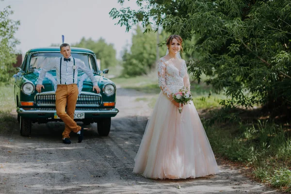 Just married wedding couple is standing near the retro vintage car in the park. Summer sunny day in forest. bride in elegant white dress with bouquet and elegant groom. Bride and groom posing by a — Stock Photo, Image