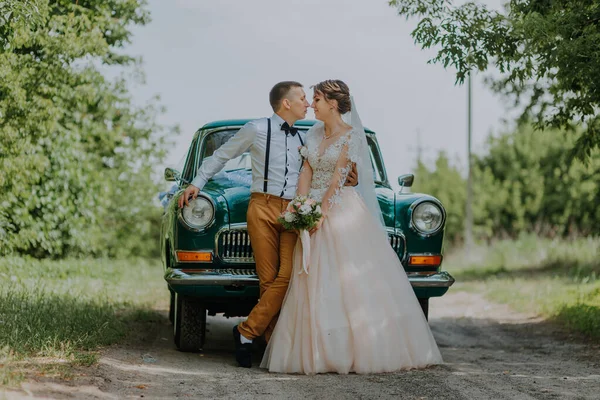 Appena sposati coppia di nozze è in piedi vicino alla macchina d'epoca retrò nel parco. Estate giornata di sole nella foresta. sposa in elegante abito bianco con bouquet ed elegante sposo innamorato abbraccio. Sposa e sposo — Foto Stock