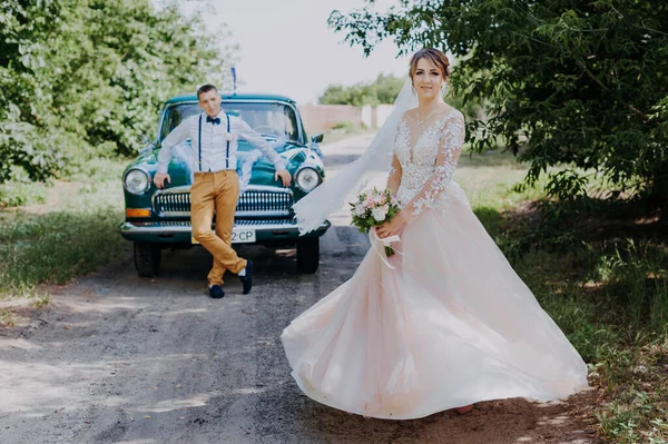Just married wedding couple is standing near the retro vintage car in the park. Summer sunny day in forest. bride in elegant white dress with bouquet and elegant groom. Bride and groom posing by a — Stock Photo, Image