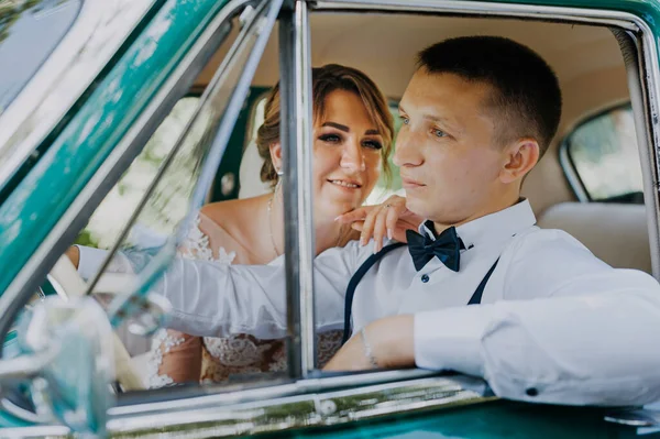 Snygg snygg blond brud poserar i retro grön bil med brudgum. Bruden och brudgummen sitter inne i retrobilen. Bröllop, brud och brudgum med en veteranbil — Stockfoto