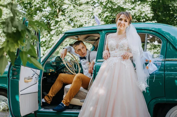 Just married wedding couple is standing near the retro vintage car in the park. Summer sunny day in forest. bride in elegant white dress with bouquet and elegant groom in love hugging. Bride and groom — Stock Photo, Image