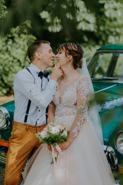 Casamento casal apenas está de pé perto do carro vintage retro no parque. Verão dia ensolarado na floresta. noiva em vestido branco elegante com buquê e noivo elegante no abraço do amor. Noiva e noivo — Fotografia de Stock