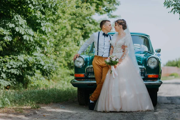 Appena sposati coppia di nozze è in piedi vicino alla macchina d'epoca retrò nel parco. Estate giornata di sole nella foresta. sposa in elegante abito bianco con bouquet ed elegante sposo innamorato abbraccio. Sposa e sposo — Foto Stock