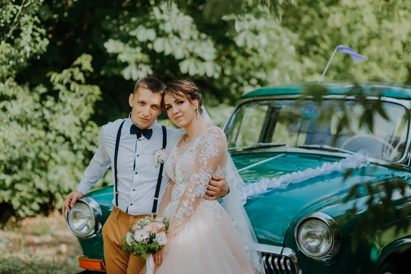 Yeni evli bir çift parktaki antika arabanın yanında duruyor. Ormanda güneşli bir yaz günü. Şık, beyaz elbiseli, buketli gelin ve aşık damat. Gelin ve damat — Stok fotoğraf