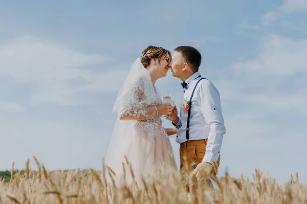 Жених и невеста держат бокалы свадебного шампанского на фоне пшеничного поля. Счастливой свадебной пары на пшеничном поле. Невеста в белом платье и груме весело проводит летний день. Только что женился. — стоковое фото