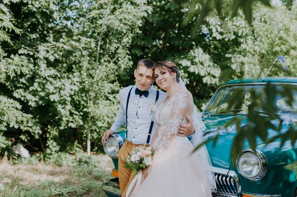 Yeni evli bir çift parktaki antika arabanın yanında duruyor. Ormanda güneşli bir yaz günü. Şık, beyaz elbiseli, buketli gelin ve aşık damat. Gelin ve damat — Stok fotoğraf