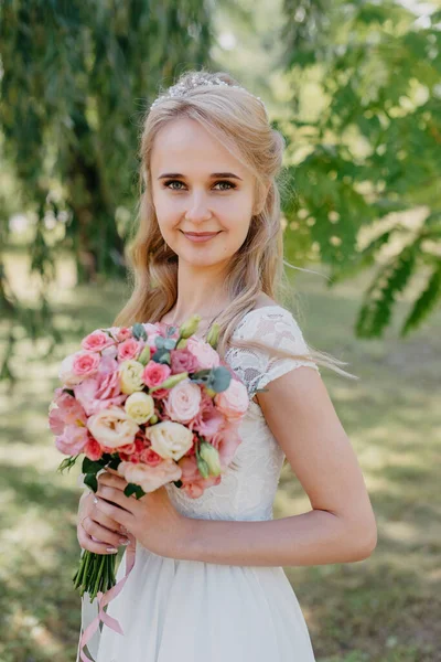 Açık havada çiçekli gelin. Orman parkında bir buket çiçekle uzun beyaz gelinlik içinde güzel bir gelinin portresi. Moda gelinliği içinde, doğal bir geçmişi olan güzel bir gelin.