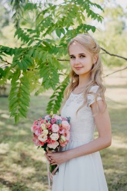 Açık havada çiçekli gelin. Orman parkında bir buket çiçekle uzun beyaz gelinlik içinde güzel bir gelinin portresi. Moda gelinliği içinde, doğal bir geçmişi olan güzel bir gelin.