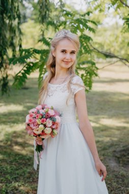 Açık havada çiçekli gelin. Orman parkında bir buket çiçekle uzun beyaz gelinlik içinde güzel bir gelinin portresi. Moda gelinliği içinde, doğal bir geçmişi olan güzel bir gelin.