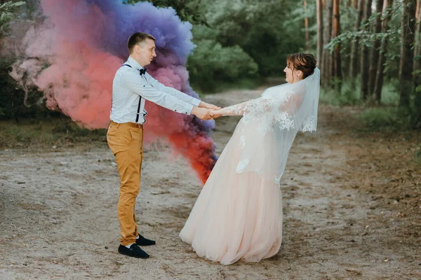 Bride and groom with the blue and yellow colored smoke bombs in the summer park. The bride and groom with the colored smoke at the background of summer forest. Wedding couple runs color smoke in the — Fotografia de Stock