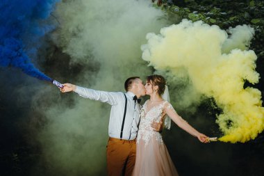 Gelin ve damat, yaz parkında mavi ve sarı renkli sis bombalarıyla. Gelin ve damat, yaz ormanının arka planında renkli dumanlarla. Düğün çifti renkli sigara içiyor.