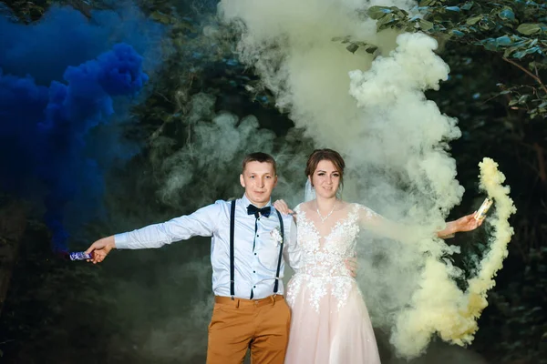 Noiva e noivo com as bombas de fumaça coloridas azuis e amarelas no parque de verão. A noiva e o noivo com a fumaça colorida no fundo da floresta de verão. Casamento casal corre fumaça cor no — Fotografia de Stock