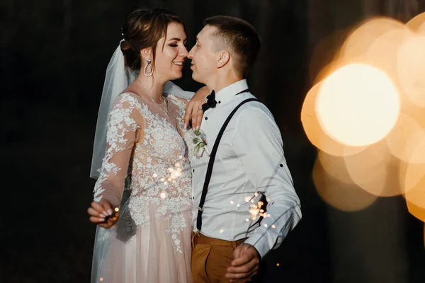 Newlyweds with sparklers in a night park. The newlyweds kiss and hold the sparkler. A perfect end to the wedding day. shooting in the dark. the bride in a white elegant dress and the groom in a white — Stockfoto
