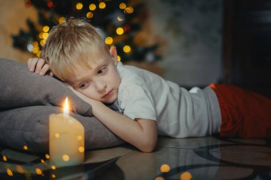 5-7 yaşlarında gülümseyen sevimli bir çocuğun portresi bir Noel ağacının arka planında yerde yatıyor. Seçici yumuşak odaklanma. Noel ağacının yanında yerde yatan sarışın çocuk. Yeni Yıl