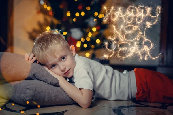 Portrait of a smiling cute boy 5-7 years old lying on the floor against the background of a Christmas tree. Selective soft focus. blond boy near the Christmas tree lying on the floor. New Year — Stock Photo, Image