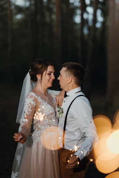 Recém-casados com faíscas em um parque noturno. Os recém-casados beijam e seguram o sparkler. Um fim perfeito para o dia do casamento. a disparar no escuro. a noiva em um vestido elegante branco e o noivo em um branco — Fotografia de Stock