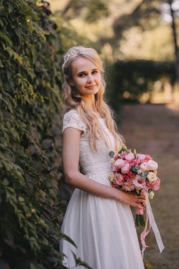 Açık havada çiçekli gelin. Orman parkında bir buket çiçekle uzun beyaz gelinlik içinde güzel bir gelinin portresi. Moda gelinliği içinde, doğal bir geçmişi olan güzel bir gelin.