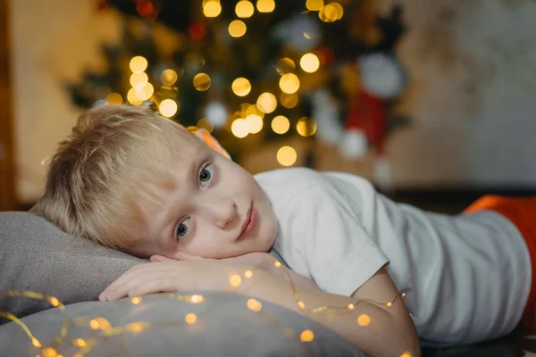 Portrait of a smiling cute boy 5-7 years old lying on the floor against the background of a Christmas tree. Selective soft focus. blond boy near the Christmas tree lying on the floor. New Year — Stock Photo, Image