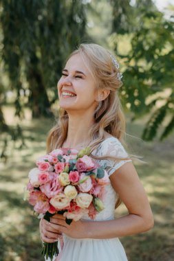 Açık havada çiçekli gelin. Orman parkında bir buket çiçekle uzun beyaz gelinlik içinde güzel bir gelinin portresi. Moda gelinliği içinde, doğal bir geçmişi olan güzel bir gelin.