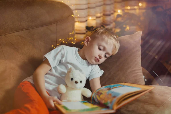 3-4 years old boy is reading a book sitting on a sofa with teddy bear. Home reading of fairy tales. Cute little boy is reading book while sitting on a sofa. Child reads a book to his best bear friend. — Stockfoto