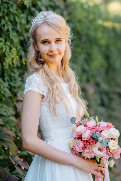 Sposa con fiori all'aperto. Ritratto di una bella sposa in un lungo abito da sposa bianco con un mazzo di fiori in un parco forestale. Bella sposa in abito da sposa di moda su sfondo naturale.Il — Foto Stock