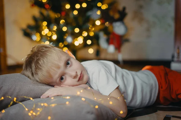 Portrait of a smiling cute boy 5-7 years old lying on the floor against the background of a Christmas tree. Selective soft focus. blond boy near the Christmas tree lying on the floor. New Year — Stock Photo, Image