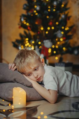 5-7 yaşlarında gülümseyen sevimli bir çocuğun portresi bir Noel ağacının arka planında yerde yatıyor. Seçici yumuşak odaklanma. Noel ağacının yanında yerde yatan sarışın çocuk. Yeni Yıl