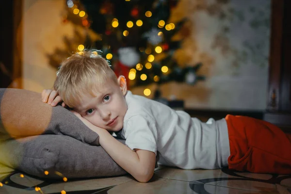 Portrait of a smiling cute boy 5-7 years old lying on the floor against the background of a Christmas tree. Selective soft focus. blond boy near the Christmas tree lying on the floor. New Year — Stock Photo, Image
